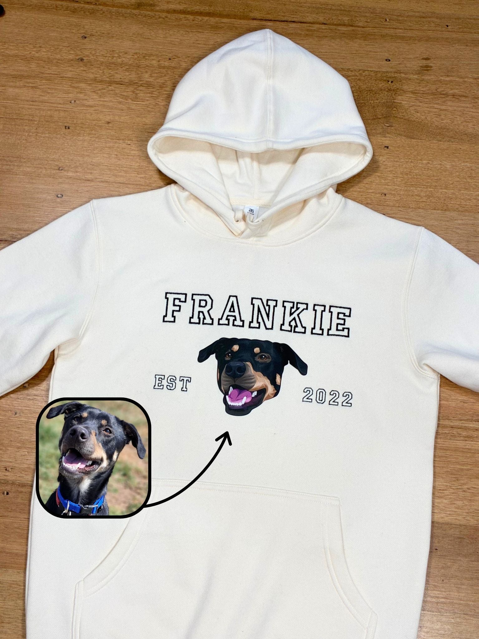 White hoodie with a dog's face and name 'Frankie' printed on it, on a wooden surface.
