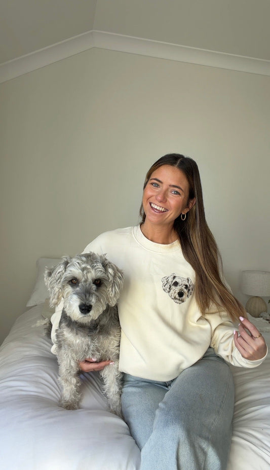Woman sitting on a bed with a dog, wearing a cream sweatshirt with a dog print.